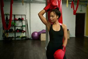 retrato do lindo africano Esportes mulher, atraente fêmea boxer dentro Preto roupa de esporte e vermelho boxe luvas, olhando para a lado, inclinado em uma soco saco às boxe academia. ativo estilos de vida conceito foto