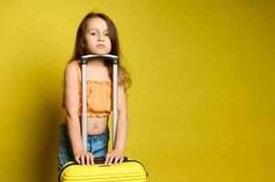 lindo pequeno menina dentro à moda verão laranja topo e casual azul jeans, em pé perto uma vibrante na moda amarelo mala de viagem foto