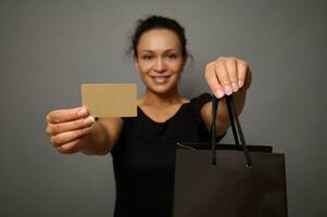 foco em em branco ouro plástico crédito ou desconto cartão e Preto compras saco dentro a mãos do borrado bonita mulher com lindo sorriso, isolado sobre cinzento fundo com cópia de espaço para Preto Sexta-feira de Anúncios foto