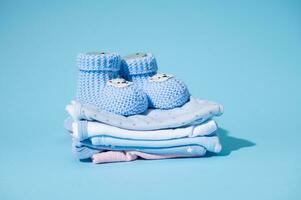 fofa azul tricotado bebê chuteiras em uma pilha do limpar \ limpo lavado passado a ferro recém-nascido bodysuits, isolado sobre azul pastel fundo foto