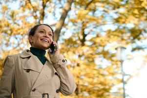 lindo alegre africano americano mulher, bem sucedido o negócio mulher dentro casual vestuário fala em Móvel telefone sorrisos cheio de dentes sorrir em a outonal parque fundo. comunicação, conexão conceito foto