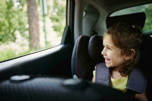 adorável bebê menina goza a viagem de carro, parece Fora do a aberto janela enquanto sentado dentro uma segurança reforço carro assento foto