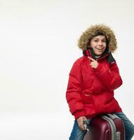 bonito europeu Garoto vestido dentro brilhante vermelho parka segurando avião brinquedo modelo e apontando em uma cópia de espaço do branco fundo enquanto sentado em dele bagagem. inverno feriados viagem, turismo conceito foto