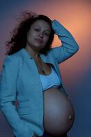 lindo multiétnico grávida mulher, olhando às Câmera, posando com nu barriga em estúdio fundo com néon luzes foto