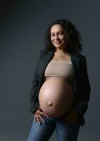 blinda jovem adulto grávida mulher com grande barriga dentro gravidez trimestre terceiro, sorridente às Câmera, isolado em cinzento foto