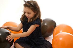 adorável bebê garota, 4 anos velho criança vestindo a tarde vestir e aro com gato ouvidos, tendo Diversão jogando com brilhante laranja e Preto balões em branco fundo com cópia de espaço. dia das Bruxas conceito foto