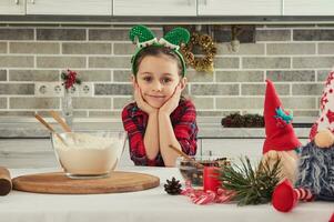 retrato do uma lindo adorável europeu pequeno menina com duende aro em dela cabeça fofinho sorrisos olhando às Câmera sentado às uma cozinha mesa com ingredientes para cozinhando uma Natal bolo foto