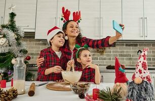 alegre família do lindo mãe dentro santa chapéu e dela adorável crianças, filho e filha falando de vídeo chamar, fazer selfie, gravação vídeo, cozinhando Natal pão juntos dentro a casa cozinha foto