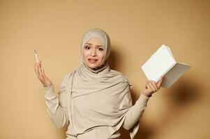 muçulmano mulher expressando surpresa, insatisfação em pé contra bege fundo com bloco de anotações e lápis dentro mãos foto