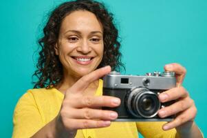 alegre africano americano mulher fotógrafo dentro brilhante amarelo roupas sorridente cheio de dentes sorrir enquanto segurando a velho retro estilo vintage Câmera e fotografando, levando As fotos contra azul fundo