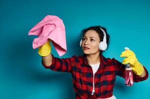 mulher dentro fones de ouvido e amarelo borracha luvas detém detergente spray e limpeza pano, simulando limpeza superfície. foto