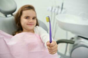 foco em uma escova de dente dentro a mão do uma alegre bonita menina fofinho sorridente olhando às Câmera sentado em Dentistas cadeira durante dental checar. oral Cuidado e higiene. pediatra odontologia conceito foto