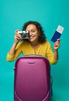 alegre sorridente hispânico mulher segurando uma Passaporte com bilhete e embarque passar e uma vintage Câmera, fotografando, sorridente cheio de dentes sorrir olhando às Câmera contra azul colori fundo. cópia de espaço foto