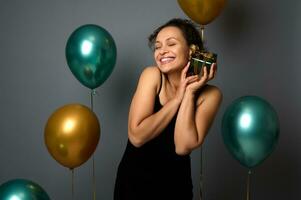 fofa mulher com fechadas olhos suavemente abraços dela Natal presente, em pé perto lindo brilhante ar balões do verde metálico e dourado cores, isolado retrato em cinzento fundo com cópia de espaço para de Anúncios foto