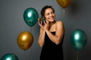 alegre misturado raça bonita mulher com lindo sorrir detém uma presente caixa perto dela face, poses em cinzento fundo com dourado verde ar balões. aniversário, Natal, Novo ano conceitos para anúncio. foto