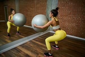 traseiro Visão do uma desportivo mulher segurando uma cinzento ginástica bola e fazendo glúteo exercícios, bombeamento nádegas enquanto agachado dentro frente do uma espelho dentro ginástica estúdio foto