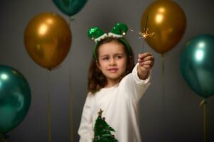 lindo bebê menina vestindo duende aro e caloroso branco acolhedor suéter com brilhar Natal árvore carrinhos contra brilhante ouro verde inflado ar balões e ilumina estrelinhas a comemorar a 2022 Novo ano foto