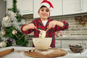 bonito pré-adolescente caucasiano garoto, adorável criança chefe de cozinha blogueiro dentro santa chapéu amassar massa e mostrando isto para a Câmera, fofinho sorridente com alegre cheio de dentes sorriso. Natal culinária conceito foto