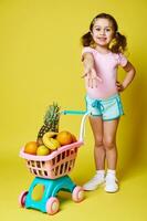 lindo pequeno menina dentro verão roupas apontando dela mão, mostrando uma compras carrinho cheio do delicioso frutas. isolado em amarelo fundo, cópia de espaço foto