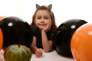 tradicional evento, dia das Bruxas festa conceito. lindo pequeno bruxa menina dentro aro com gato orelhas deitado em branco fundo com cópia de espaço Próximo para abóbora e laranja Preto balões foto