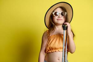 caucasiano adorável pequeno menina dentro oculos de sol, Palha chapéu e laranja principal, posando com portátil bagagem, isolado em amarelo foto