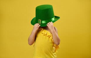 pequeno menina esconde dela face debaixo uma verde irlandês duende chapéu. santo patrick's dia conceito em amarelo fundo. foto