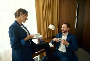 bonito jovem homem de negocios sentado em a poltrona e rindo enquanto falando para dele bonito parceiro sobre uma copo do café dentro uma hotel quarto durante uma o negócio viagem. o negócio pessoas em café pausa foto