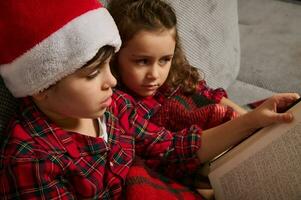 fechar-se do adorável caucasiano crianças, irmão e irmã, Garoto dentro santa chapéu e lindo menina sentado em a sofá e lendo fada conto juntos em Natal noite. foto