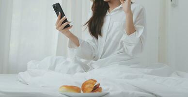 feliz jovem fêmea dentro casual roupas sorridente e olhando às Câmera enquanto usando computador portátil em cama dentro quarto às casa foto