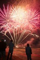 dois pessoas em pé em uma de praia assistindo uma fogos de artifício exibição foto