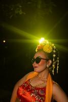 uma sundanês dançarino poses dentro frente do uma brilhando luz enquanto vestindo a laranja traje com oculos de sol e flores em dela cabeça foto