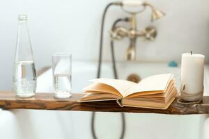uma livro, uma garrafa do água, uma vidro e uma vela em uma de madeira bandeja acima a banheiro, contra a fundo do a Antiguidade torneira foto