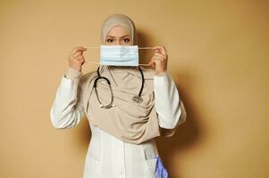 muçulmano médico com coberto cabeça dentro hijab segurando e vestindo médico mascarar em pé em bege fundo eith cópia de espaço foto