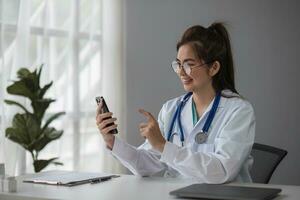 ásia fêmea médico trabalhos às hospital escritório escrivaninha dando paciente conveniência conectados serviço conselho, sorridente escrever uma prescrição ordem médico com Smartphone, saúde Cuidado, impedindo doença conceito. foto