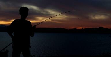 silhueta do uma pescador com uma pescaria Cajado dentro a lago às pôr do sol foto