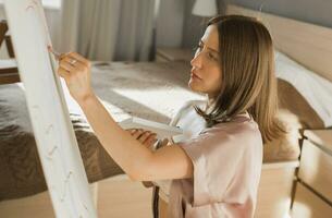jovem mulher artista pintura em tela de pintura em a cavalete às casa dentro quarto - arte e criatividade conceito foto