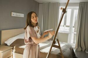 jovem mulher artista pintura em tela de pintura em a cavalete às casa dentro quarto - arte e criatividade conceito foto
