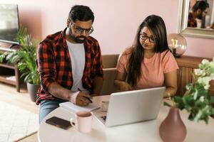 milenar indiano marido e esposa fazendo doméstico papelada, contabilidade trabalho e revendo papel contas, recibos às computador portátil computador, usando conectados calculadora - econômico crise conceito foto