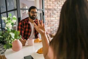 feliz casal comendo café da manhã e falando às jantar mesa dentro manhã. indiano menina e latino cara. relação e diversidade conceito foto