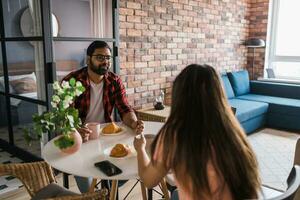 jovem diverso amoroso casal comendo croissant e fala juntos às casa dentro café da manhã tempo. comunicação e relação conceito foto