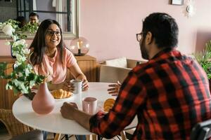 jovem diverso amoroso casal comendo croissant e fala juntos às casa dentro café da manhã tempo. comunicação e relação conceito foto