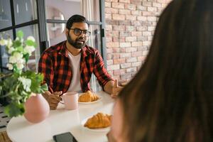 jovem diverso amoroso casal comendo croissant e fala juntos às casa dentro café da manhã tempo. comunicação e relação conceito foto