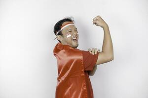 animado ásia homem vestindo uma vermelho principal, bandeira capa e arco de cabelo, mostrando Forte gesto de elevação dele braços e músculos sorridente orgulhosamente. da indonésia independência dia conceito. foto