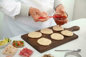 Senior mulher adicionando pizza molho para a esticado massa. delicioso caseiro mini pizzas preparação. foto