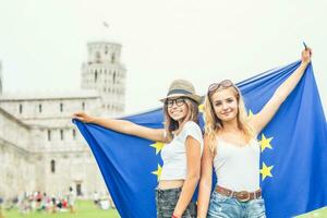 jovem adolescente meninas viajante com bandeira do europeu União antes a histórico torre dentro Cidade pisa - Itália foto