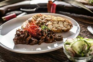 porcelana prato cheio do veado carne de veado Goulash ensopado servido com bolinho de massa e fresco salada Como a Aperitivo. Comida é cercado de Caçando acessórios tal Como Caçando espingarda, balas e rústico faca foto