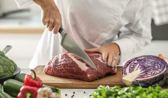 fêmea chefe de cozinha cortes através cru carne carne em uma de madeira corte borda cercado de legumes foto