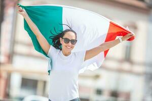 sorridente bonito menina dentro oculos de sol e branco camiseta detém italiano bandeira em uma rua a comemorar foto