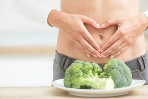 mulher com Esportes figura em dela barriga mostra coração forma. fresco brócolis dentro prato em cozinha mesa foto