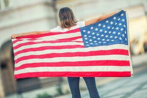 feliz jovem americano escola menina segurando e acenando dentro a cidade com EUA bandeira foto